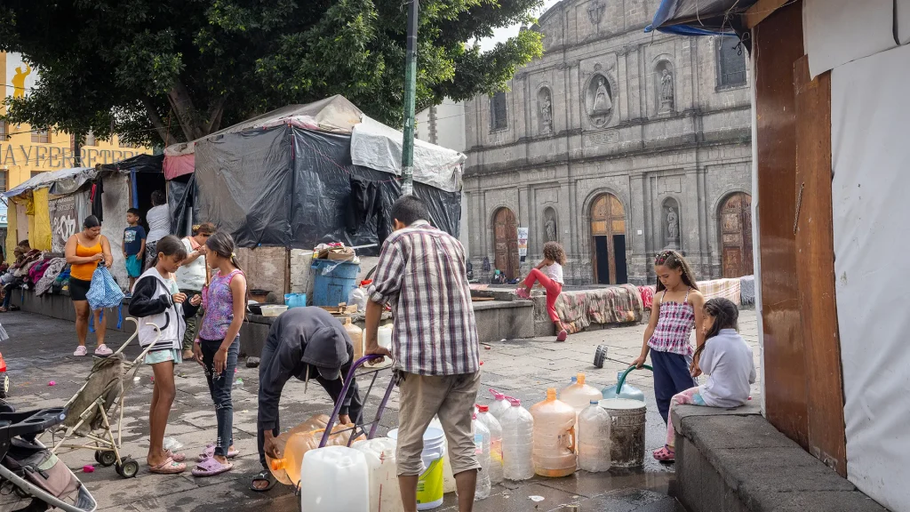 Personas migrantes atrapados entre la violencia, las estrategias de disuasión y los asfixiantes procedimientos de asilo. Un grupo de niños y adultos recoge agua en la Plaza de la Soledad, en la Ciudad de México.