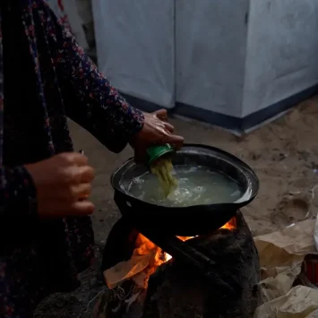 Una madre prepara comida para sus hijos y familia, vertiendo una lata de judías verdes en agua hirviendo sobre un fuego que ella misma enciende. La cantidad de ayuda humanitaria que entra en Gaza alcanza su nivel más bajo en meses.