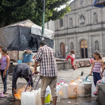 Personas migrantes atrapados entre la violencia, las estrategias de disuasión y los asfixiantes procedimientos de asilo. Un grupo de niños y adultos recoge agua en la Plaza de la Soledad, en la Ciudad de México.