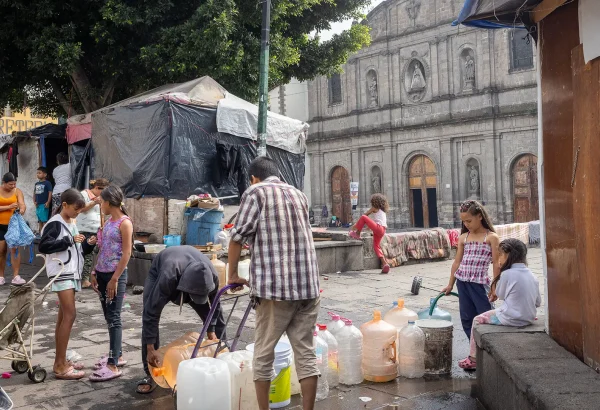 Personas migrantes atrapados entre la violencia, las estrategias de disuasión y los asfixiantes procedimientos de asilo. Un grupo de niños y adultos recoge agua en la Plaza de la Soledad, en la Ciudad de México.
